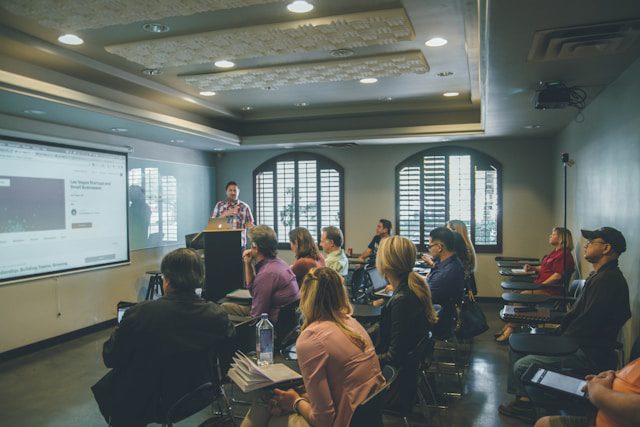 Speaker giving a presentation in a classroom-style setting with attendees seated and viewing a projected screen.